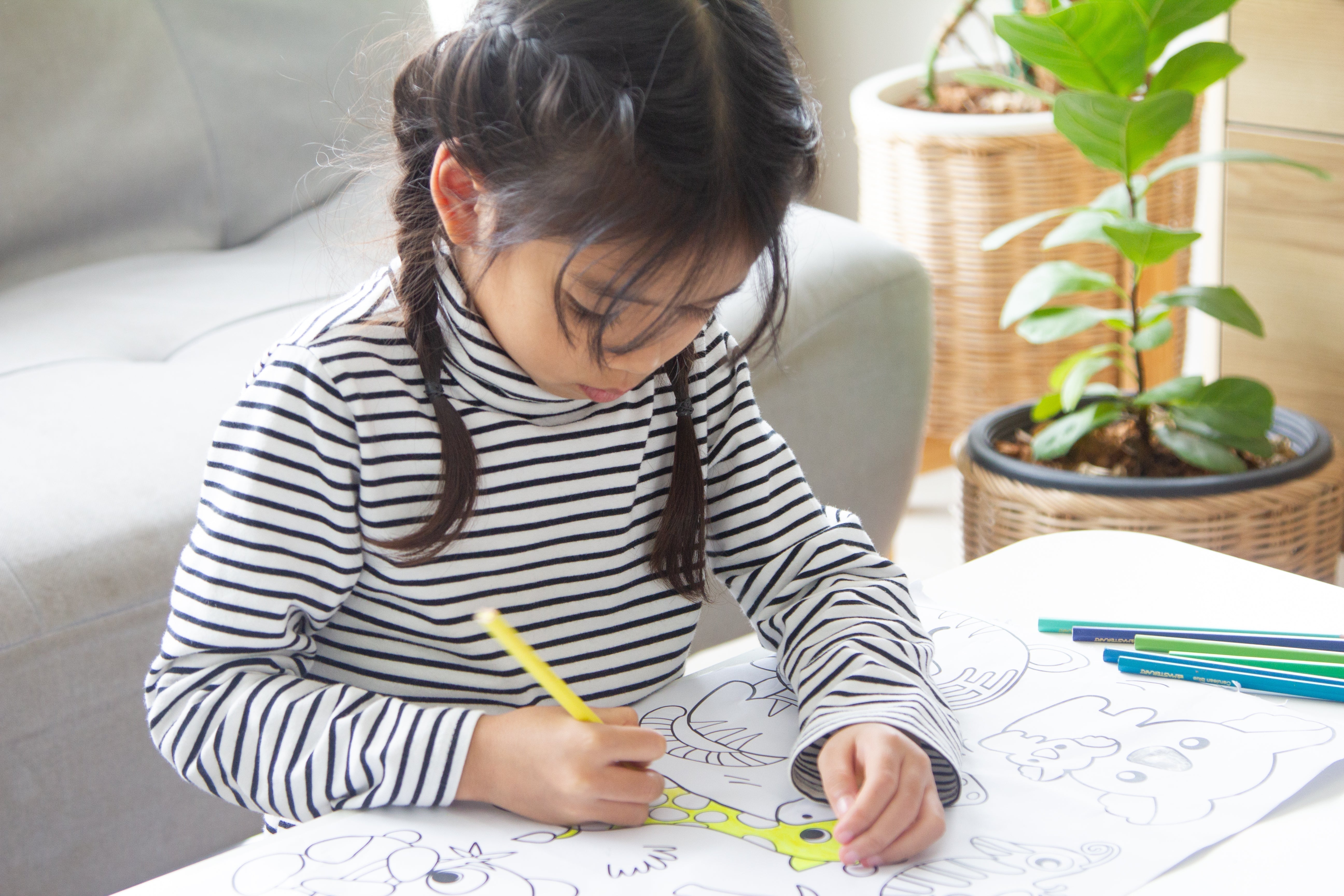 Young girl with dark braids in a striped turtleneck colors a giraffe in a colouring book at a white table, using a yellow pencil beside scattered pencils, leaning forward in focus, with a grey sofa and plant behind