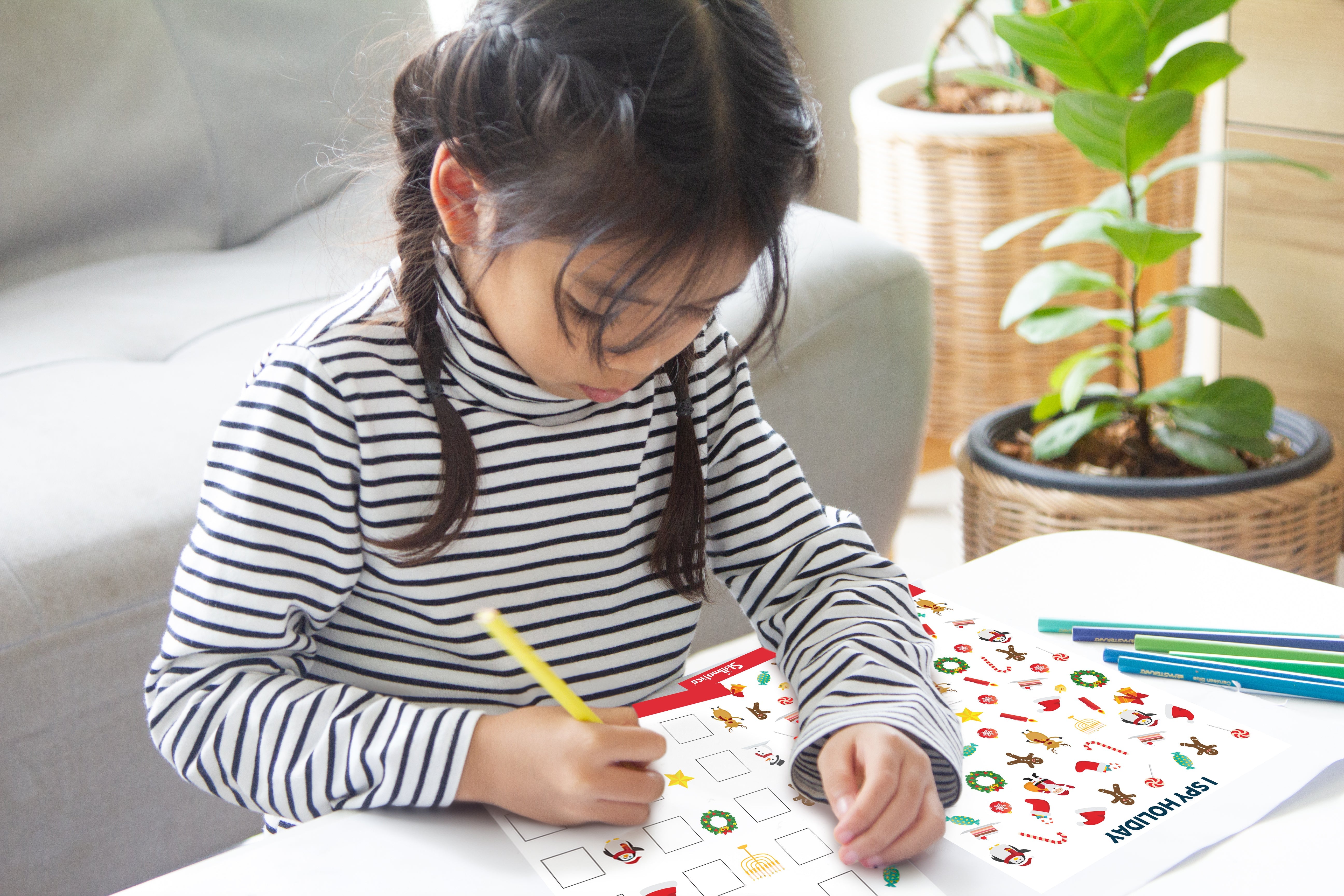 Young girl with dark braided hair in a black-and-white striped turtleneck works on a holiday-themed activity book at a white table, holding a yellow pencil, focused, with coloured pencils nearby and a sofa and potted plants behind