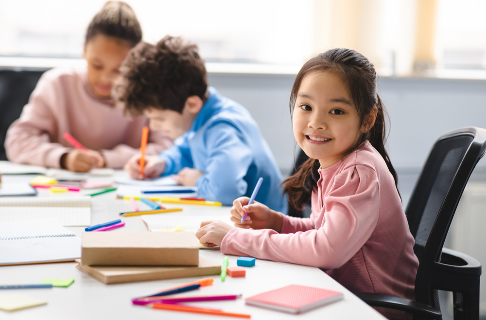 Three kids doing art & craft activity on a white table. One girl in pink tshirt is looking into the camera and smiling