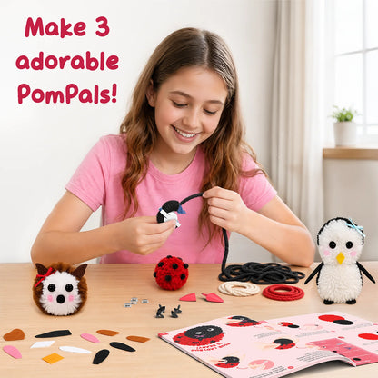 A child in a pink T-shirt uses the Skillmatics PomPals maker to wrap black yarn while making pom-pom animals at a table. Finished hedgehog, penguin and ladybug crafts sit nearby, alongside yarn, felt shapes, craft accessories and an illustrated instruction guide.