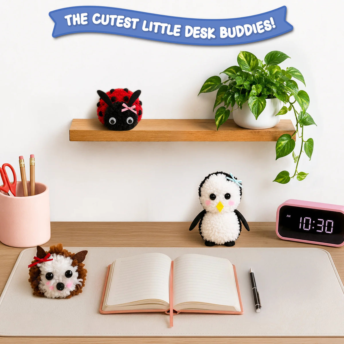 Three finished Skillmatics PomPals yarn animals—a red-and-black ladybug, a white penguin and a brown-and-white hedgehog—are displayed on a desk and shelf beside a notebook, pen, clock, stationery holder and potted plant, showing them as cute desk companions.