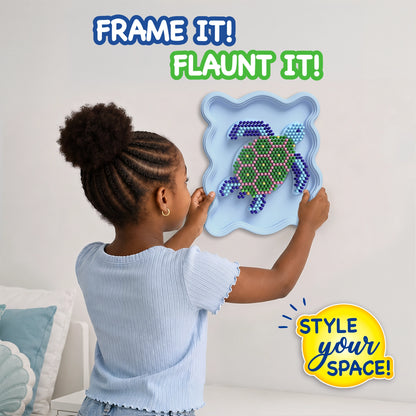 Child placing a completed Skillmatics Stixels Sea Turtle artwork onto a bedroom wall. The framed 3D wall decor features colorful Stixels arranged on a turtle-shaped design inside a light blue frame. The image showcases the finished DIY craft as personalized room décor for kids.