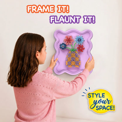 Child wearing a pink sweater hangs a completed Skillmatics Stixels Flower Vase artwork on a wall. The finished 3D design features colorful flowers and a patterned vase mounted inside a lavender decorative frame, demonstrating how the craft transforms into personalized wall décor for a bedroom or playroom.